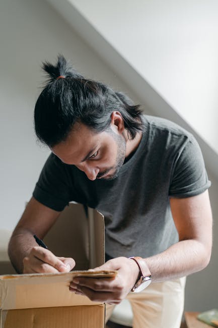 A man with dark hair tied back into a small ponytail, wearing a grey T-shirt, is inside a home, packing a cardboard box with various items. He is leaning over a partially filled box on a surface, carefully folding or sealing the flaps. In the background, there is natural daylight coming through a window, casting soft shadows on the wall. This scene depicts the packing process during home relocation, with the focus on moving preparations carried out by Man with Van Hatch End, a professional removals provider. The environment appears clean and organized, highlighting typical packing materials such as cardboard boxes and paper or plastic wrapping visible nearby, emphasizing the logistics involved in furniture transport and house removals in the Hatch End area.