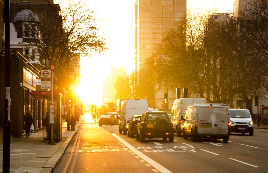 The image depicts a busy urban street during sunset in Hatch End, with a row of parked white and dark-colored vans and cars along the kerbside, some of which are engaged in a home relocation or furniture transport process. A man with a van service from Man with Van Hatch End is visible near a large white van, possibly involved in loading or unloading furnishings. The scene includes a pedestrian bus stop with a glass shelter, several pedestrians walking on the sidewalk, and a bus stop sign, all set against a backdrop of modern high-rise buildings, leafless trees, and a golden, glowing sky that casts long shadows across the street. The environment appears to be within an area designated for vehicle loading and passenger transportation, with clear markings on the pavement indicating loading zones. The overall scene captures the logistical aspects of house removals and packing and moving activities within a city setting, emphasizing the importance of adhering to local permit rules for van loading, as referenced in the Harrow Council permit regulations for Hatch End.