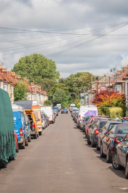 The image depicts a busy urban street during sunset in Hatch End, with a row of parked white and dark-colored vans and cars along the kerbside, some of which are engaged in a home relocation or furniture transport process. A man with a van service from Man with Van Hatch End is visible near a large white van, possibly involved in loading or unloading furnishings. The scene includes a pedestrian bus stop with a glass shelter, several pedestrians walking on the sidewalk, and a bus stop sign, all set against a backdrop of modern high-rise buildings, leafless trees, and a golden, glowing sky that casts long shadows across the street. The environment appears to be within an area designated for vehicle loading and passenger transportation, with clear markings on the pavement indicating loading zones. The overall scene captures the logistical aspects of house removals and packing and moving activities within a city setting, emphasizing the importance of adhering to local permit rules for van loading, as referenced in the Harrow Council permit regulations for Hatch End.
