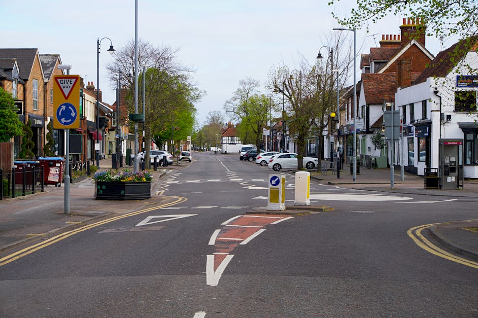 A quiet urban street in Hatch End with a narrow central road marked by white dashed lines and a small, rectangular red and brown traffic island in the foreground. The island is bounded by white dashed lines and has blue and white directional arrows, with yellow double lines running along the edges of the street. On the left side, there are residential buildings with brick and white facades, some with small front gardens bordered by black metal railings, and a large black traffic sign indicating 'GIVE WAY' with a roundabout symbol beneath. Several small trees with fresh green leaves are planted along the pavement, which is lined with street lamps and parked cars, including a white van behind the traffic island. On the right, there are commercial shops with signs, a white building with an ATM, and a parking meter, all set back from the road, with the sky overhead appearing overcast. The scene captures an area suitable for home relocation activities, with opportunities for furniture transport and loading process in the urban street environment, as visible from the parked vehicles and street layout. Man with Van Hatch End occasionally operates in this vicinity, providing removals services that involve careful packing, loading, and transit as part of house moving logistics.