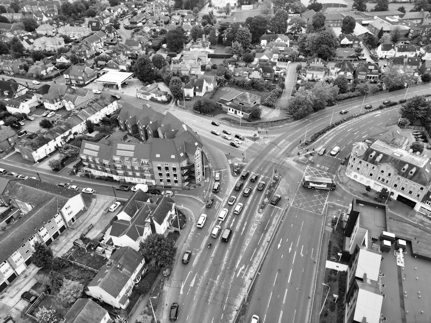 A black-and-white aerial photograph of Hatch End High Street showing a busy road with multiple lanes of traffic, including cars and trucks, some moving and others stationary. On the right side of the image, a van belonging to Man with Van Hatch End is parked in a designated loading area marked with striped lines and signage. Nearby, several cardboard boxes and wrapped furniture are arranged temporarily on the pavement, indicating an ongoing home relocation or furniture transport process. The street is lined with a variety of local shops and residential buildings, with residential houses and greenery visible further back in the background. The environment is well-lit with natural daylight, capturing the urban setting during daytime, and the image emphasizes parking, loading permits, and the logistics involved in house removals and moving services, as managed by [COMPANY_NAME].