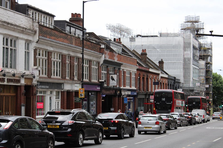 A row of multi-storey brick buildings along Hatch End High Street, with retail shops on the ground level and offices or residences above. The street scene is busy with a line of parked and moving cars, including a black Mercedes SUV, a white hatchback, and several other sedans. Two red double-decker buses are visible, one near the centre and another at the far right, with route numbers 343 and 42. To the right, a construction site is covered with scaffolding and white protective sheeting, indicating ongoing building work. Pedestrians are walking on the pavement, some approaching shop entrances. In the foreground, the edge of a residential property or shop front is visible, with a doorway and window. The overall environment suggests a typical urban high street during daytime, with natural lighting and clear weather, supporting the logistics of home relocation and furniture transport carried out by Man with Van Hatch End, who may be involved in packing, loading, or managing street loading permits for moving services.