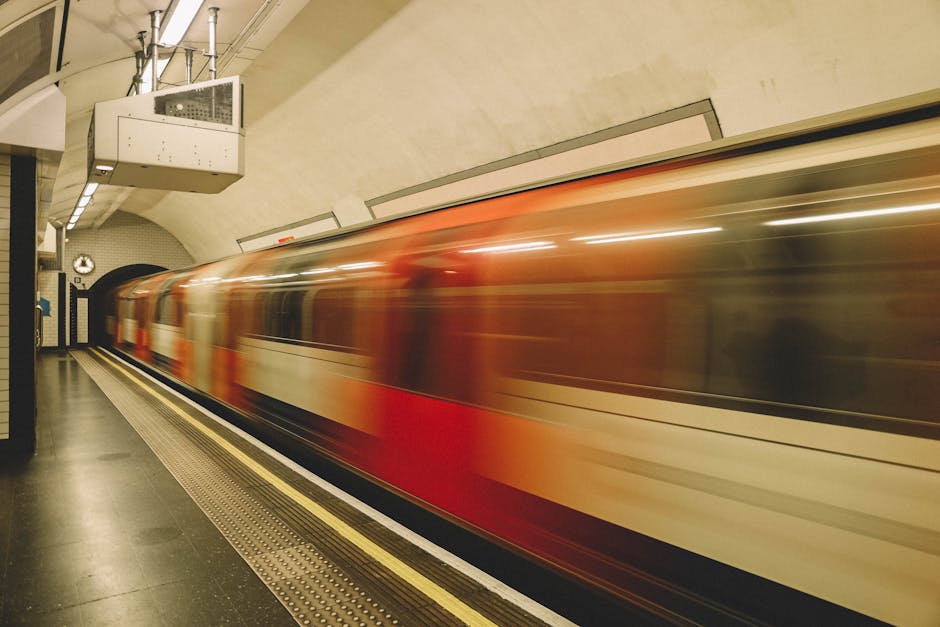 A moving train with a red and cream exterior is seen in motion, traveling through an underground station with dim lighting. The platform is visible in the foreground, featuring a tactile guiding strip along the edge for safety. The station has curved, beige-colored walls and ceiling, with overhead lighting fixtures and ventilation ducts. Behind the train, a black and white clock is mounted on the wall. The scene is captured in a long exposure, creating a blurred effect of the train's movement, which emphasizes the dynamic aspect of the home relocation process involving furniture transport and packing logistics. This image illustrates the conveyance phase of a house removal service, where efficiently coordinated transport is essential, as provided by Man with Van Hatch End, aligning with the page's focus on speeding up house sale timelines through reliable loading and transportation.