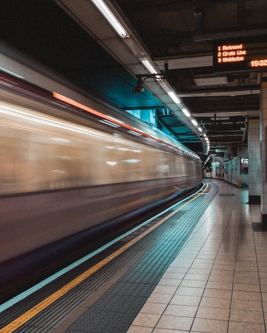 Inside an underground train station, a fast-moving train with a blurred exterior is approaching the platform. The platform features tiled flooring with tactile paving strips along the edge for safety. Overhead, illuminated digital signs display routes, including destinations such as Richmond, Circle Line, and Wimbledon. The station's ceiling includes exposed pipes and lighting fixtures, creating a utilitarian look typical of urban transit stations. On the right side, the platform curves gently, leading towards a stairway or escalator, suggesting movement towards an exit or connecting platform. This scene captures the rapid transit environment that often facilitates home relocation or furniture transport in London, with the station’s infrastructure supporting efficient luggage and moving equipment handling, an area where services such as Man with Van Hatch End operate for timely removals and local moving logistics.