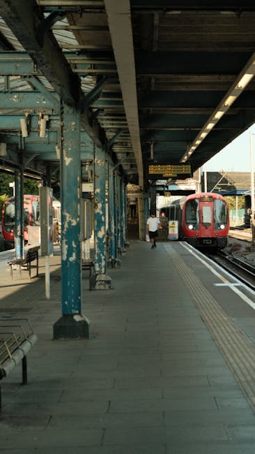 A black and white photograph shows a train station platform adjacent to railway tracks in an urban setting. Behind the platform, there is a tall brick wall with some graffitied signs and a white metal gate for access. Several street lamps and signage are visible along the platform, and a large leafless tree extends branches over the scene. In the distance, modern high-rise residential buildings rise above shorter, older structures, indicating a densely populated area. This setting captures the environment where home relocation or furniture transport services, such as those provided by Man with Van Hatch End, may operate during packing and moving processes. The image emphasizes the outdoor space involved in moving logistics, including loading and unloading stages, where boxes, furniture, or appliances might be transported via van towards or from a house or flat near the station. The overall scene highlights urban moving infrastructure relevant to efficient space-saving and residential relocations near Hatch End station.