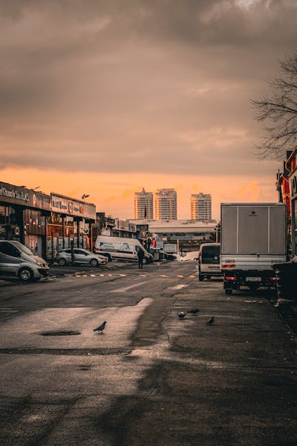 A wide view of a parking area during sunset, with a partly cloudy sky casting orange and grey hues. Several vehicles are parked, including a large white van and a box truck, both positioned at the front close to the loading zone. To the left, there are smaller cars parked near a commercial building with signage, while on the right, another vehicle is parked adjacent to a shopfront. In the background, three tall residential or office buildings are visible, with their windows reflecting the fading sunlight. The foreground features an asphalt surface with some puddles and scattered pigeons, indicating recent rain. A person is carrying a large cardboard box towards the entrance of the building, suggesting a home relocation or moving process supervised by [COMPANY_NAME], possibly involved in furniture transport or packing and moving services. The setting illustrates an urban environment prepared for a house removal, highlighting the loading process in a typical residential or commercial area.